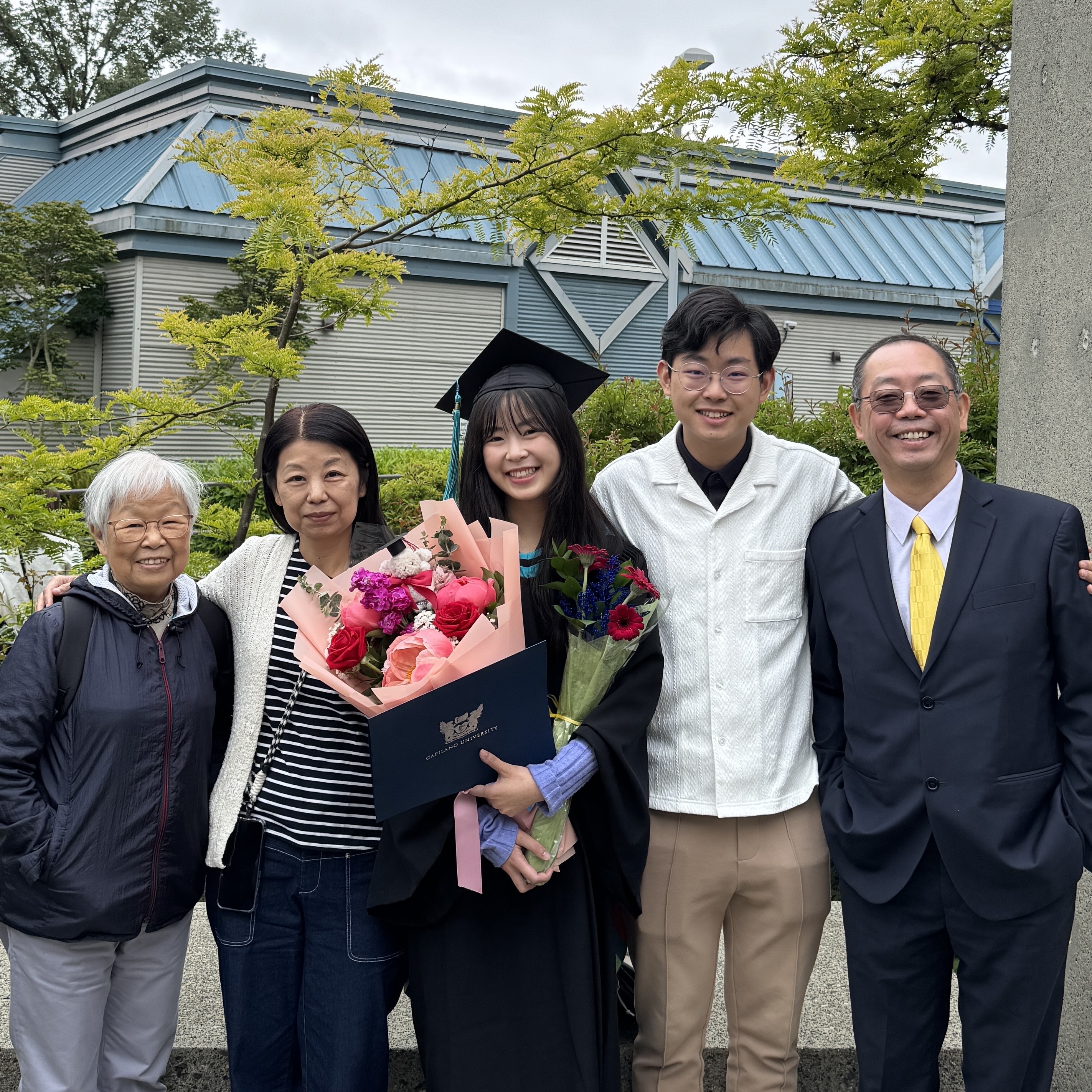 A smiling graduate in cap and gown holds flowers and a diploma, surrounded by four happy family members outdoors. The mood is joyful and celebratory.