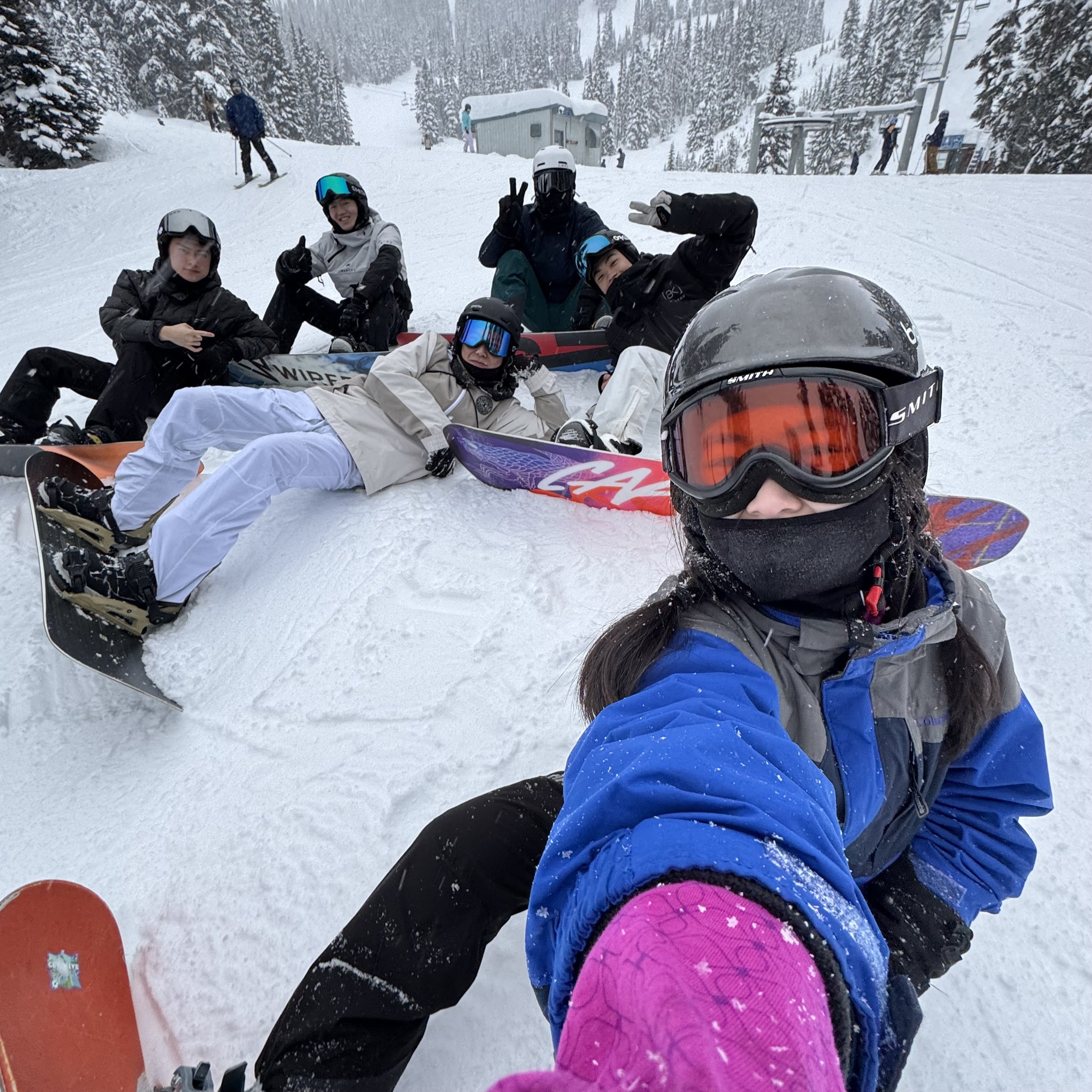 Group of six snowboarders in winter gear and helmets pose on a snowy mountain slope. They appear cheerful, with trees and a ski lift in the background.