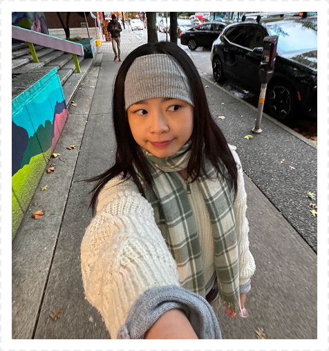 Smiling young woman with long black hair, wearing a fluffy blue cardigan and white top, sits against a plain white background. The tone is cheerful and warm.