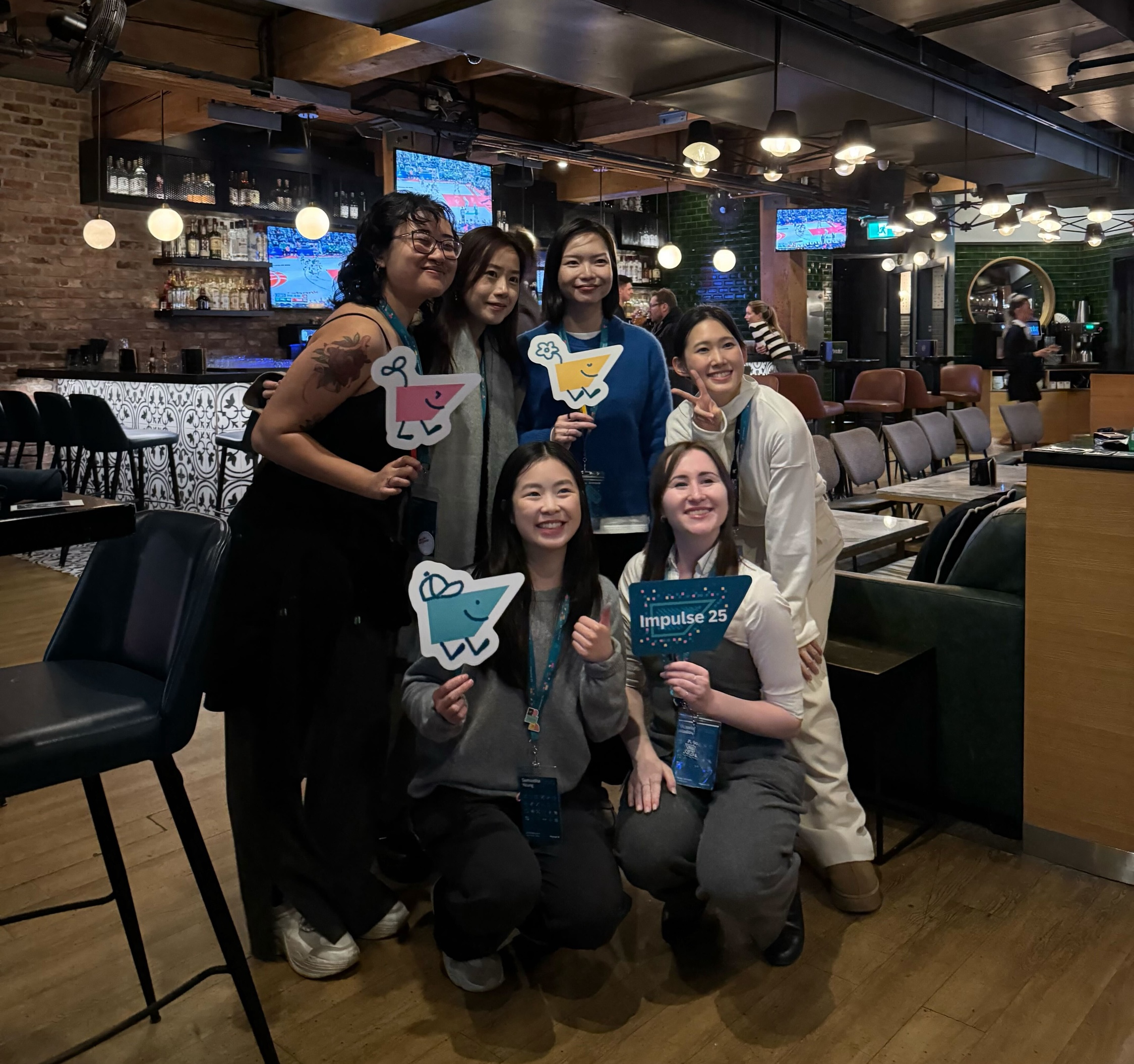 A group of seven people smiling and holding colorful cartoon cutouts and a sign reading Impulse 25. They appear cheerful and are posing playfully.