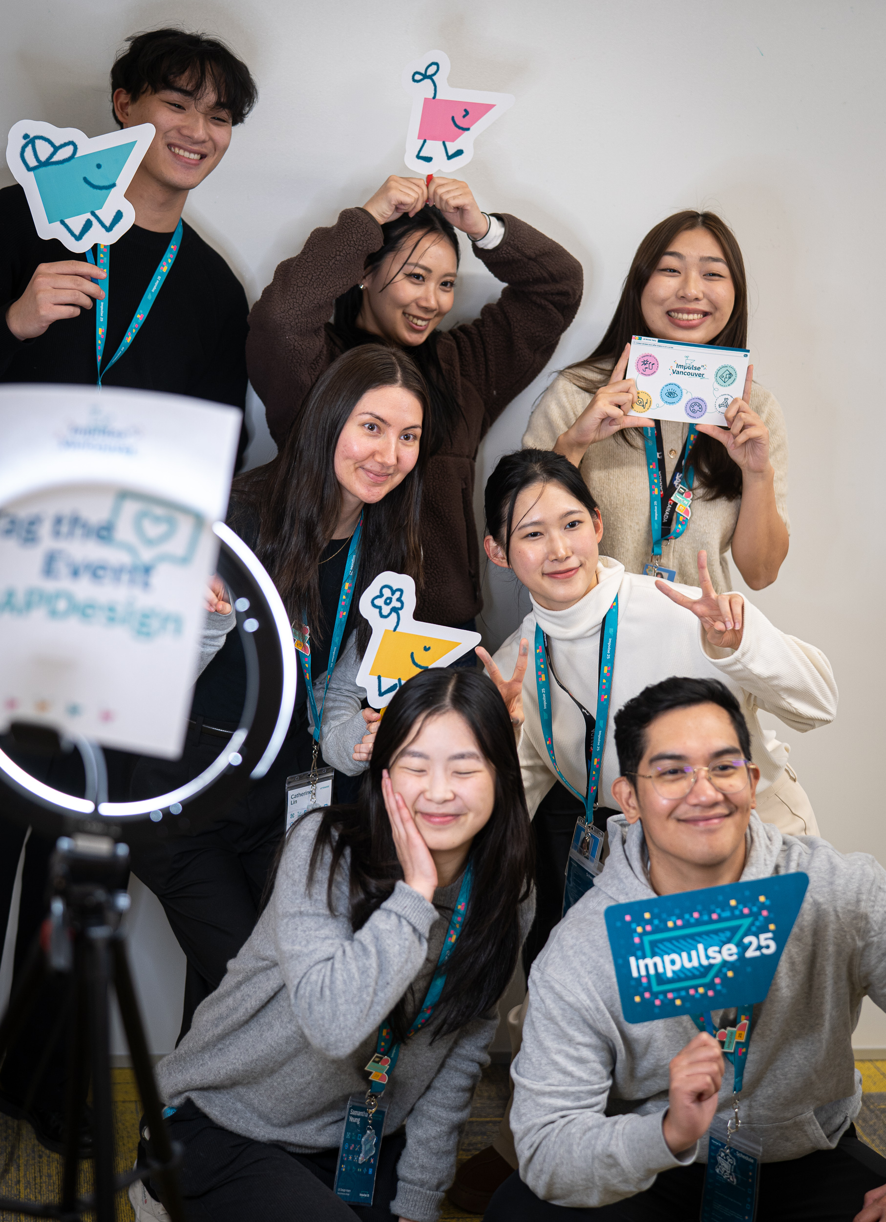 A group of seven people smiling and holding colorful cartoon cutouts and a sign reading Impulse 25. They appear cheerful and are posing playfully.